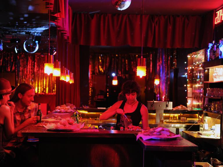 Eine Bar in Berlin, in rotes Licht getaucht. Eine Frau hinterm Tresen, zwei Menschen an der Bar.