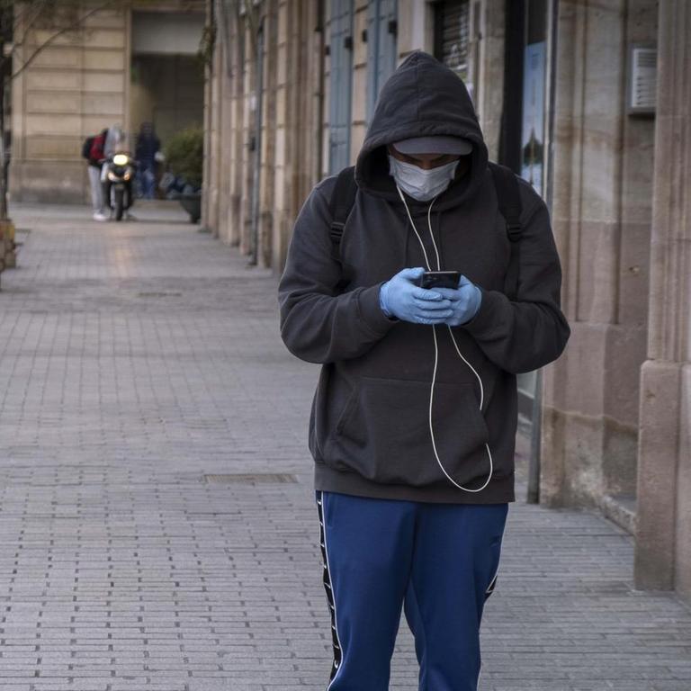 EIn mann mit Schutzmaske vor dem Mund steht auf der Straße und schaut auf sein Smartphone, April 2, 2020, Barcelona, Spanien 