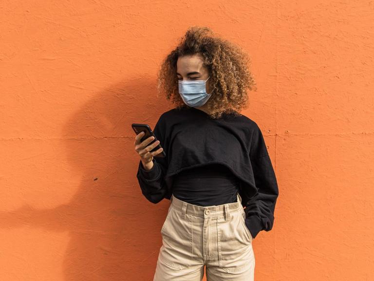 Eine Frau mit Maske steht ihr Smartphone haltend vor einer orange farbenen Wand