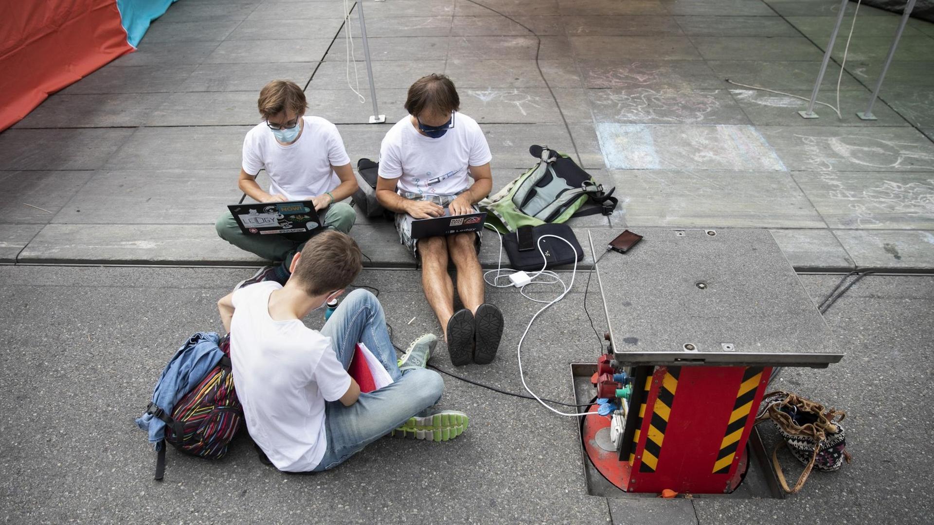 Drei junge Männer in Jeans und weißen T-Shirts sitzen mit Laptops auf der Straße.