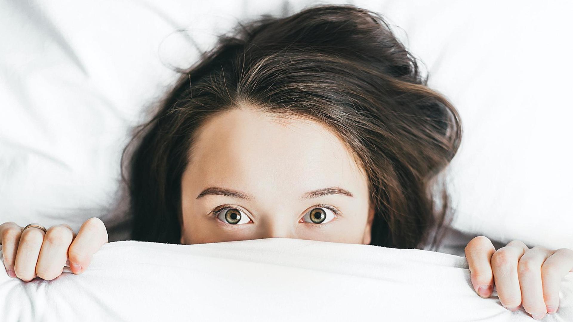 Eine Person zieht die weiße Bettdecke soweit über das Gesicht, dass nur noch die Augen und die Stirn zu erkennen sind.