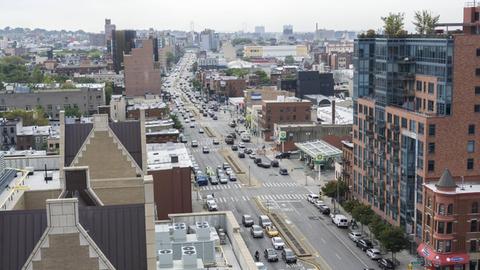 Blick von oben auf eine breite Straße im New Yorker Stadtbezirk Brooklyn. Sie wird von einigen hohen, aber auch vielen niedrigen gebäuden gesäumt und zahlreiche Autos sind unterwegs.