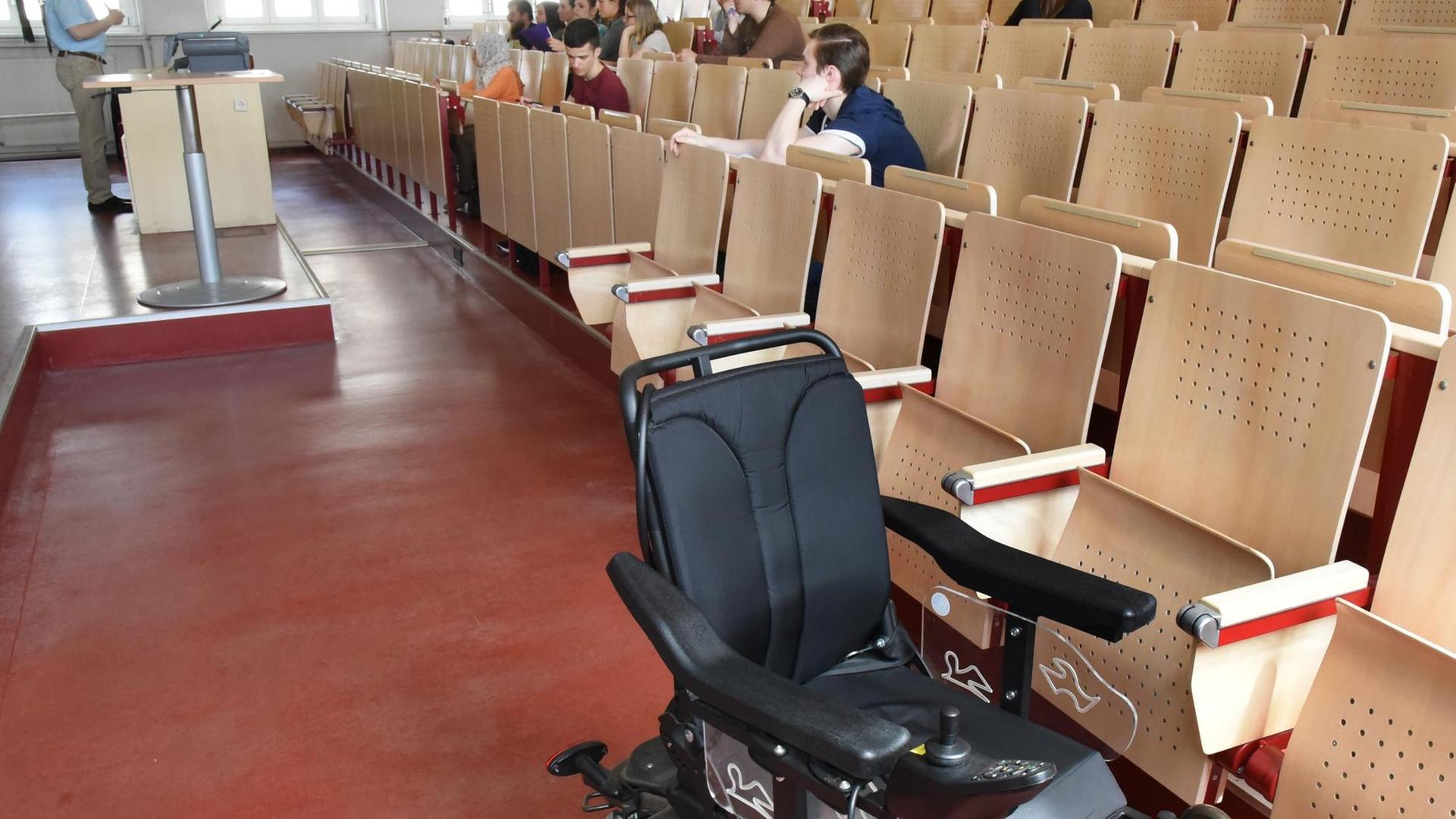 In einem Hörsaal an der Martin-Luther-Universität Halle-Wittenberg sitzen bei einer Vorlesung Studierende in den Sitzreihen. Vor der ersten Reihe steht ein leerer Rollstuhl.