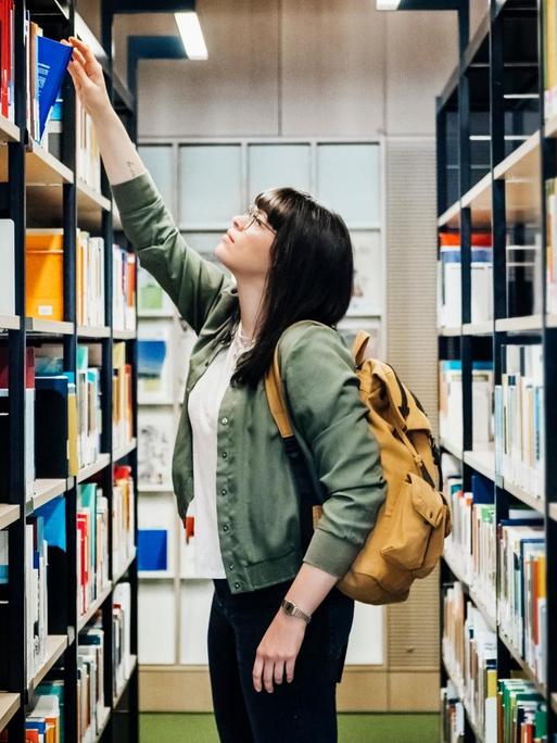 Eine junge Studentin nimmt ein Buch aus dem Regal einer Uni Bibliothek.