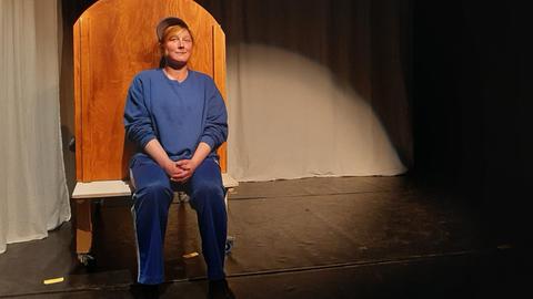 Die ehemals abhänge Schauspielerin Kathi sitzt auf einer hölzernen Bank auf einer Bühne und lächelt verschmitzt in die Kamera. Sie ist komplett in blau gekleidet und trägt eine Cap unter der ihr blonden Haare herausschauen.