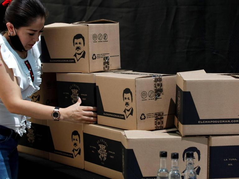 Eine Frau mit Atemschutzmaske um den Hals stapelt Kisten, die mit einer Grafik von Joaquin El Chapo Guzman versehen sind.