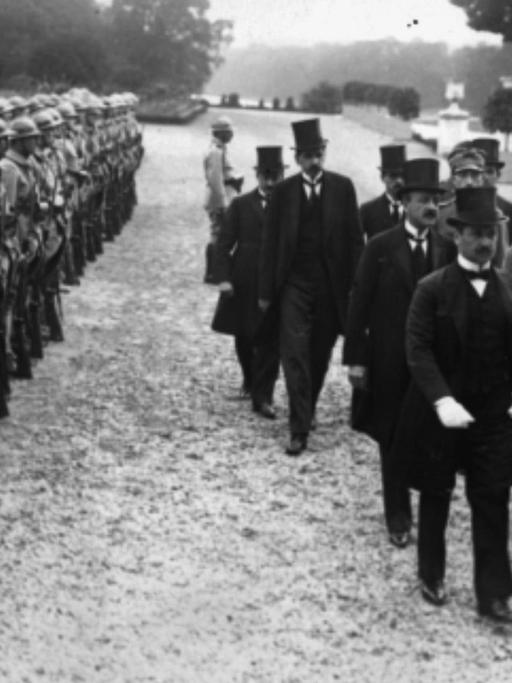 Ankunft der ungarischen Delegation, unter Führung von Dr. Benard, begleitet von Colonel Henry. Männer in Anzügen laufen an Soldatenreihen vorbei.