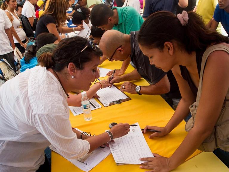 Menschen beugen sich über einen gelben Tisch, um sich in Unterschriftenlisten einzutragen.