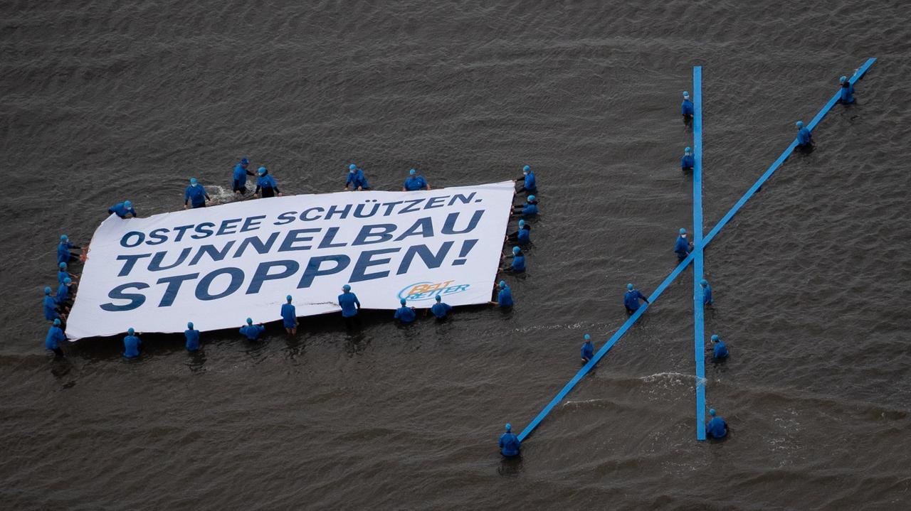 Aktivisten der Protestaktion "Beltretter" halten ein Banner mit dem Text "Ostsee schützen. Tunnelbau stoppen!", während sie im Ostseewasser stehen. 