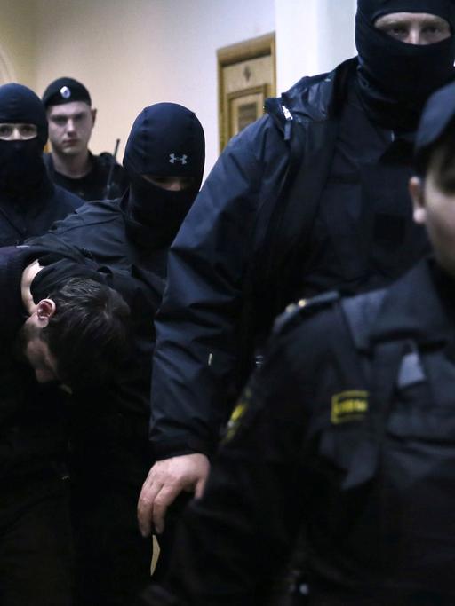 Mehrere Sicherheitskräfte in Uniform und teils vermummt begleiten einen Verdächtigen, der den Kopf gesenkt hält.