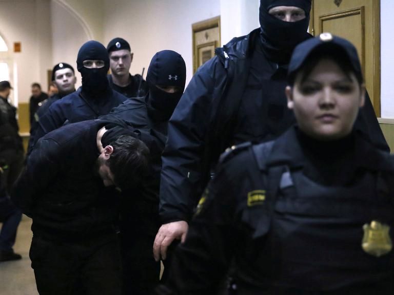 Mehrere Sicherheitskräfte in Uniform und teils vermummt begleiten einen Verdächtigen, der den Kopf gesenkt hält.