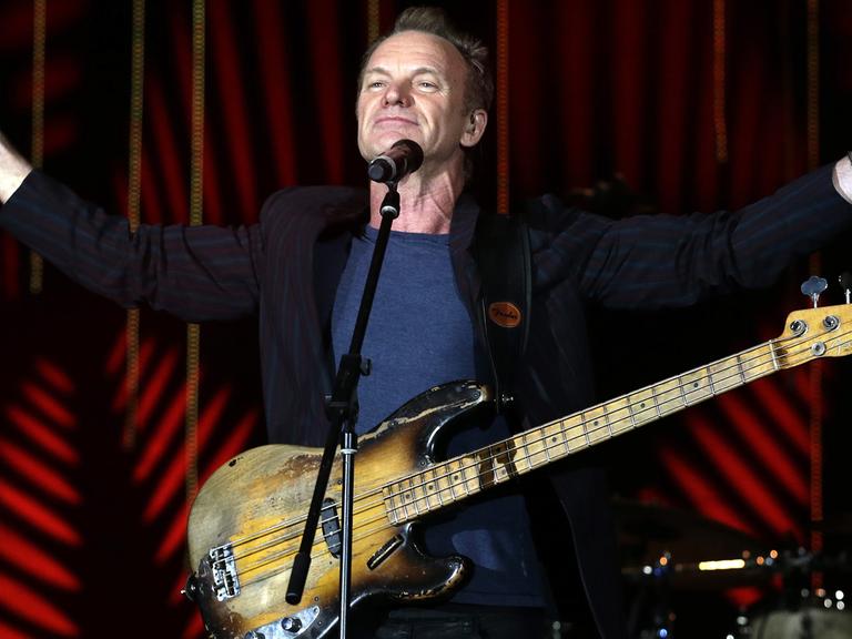 Der britische Musiker Sting steht bei einem Auftritt beim Java Jazz Festival im März 2016 in Jakarta, Indonesien mit umgehängter Gitarre vor einem Mikrofon auf der Bühne und streckt beide Arme in die Höhe.