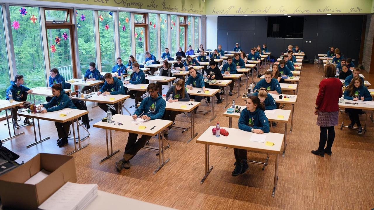 Totale des Klassenraums: Schüler sitzen an getrennten Tischen während der Abi-Prüfung, die Lehrerin führt Aufsicht