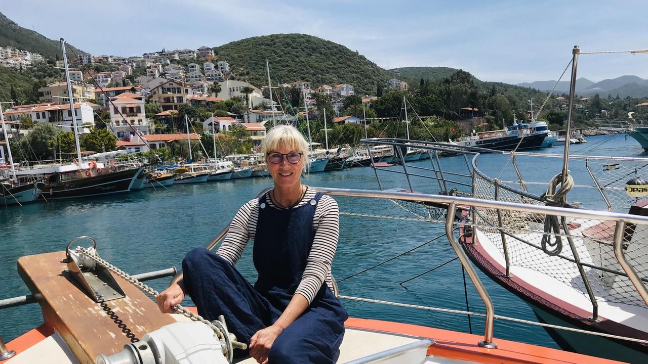 Eine blonde Frau in dunkelblauer Latzhose und mit Brille sitzt auf einem Boot. Im Hintergrund das Meer und die andere Seite des Hafens mit vielen Schiffchen.