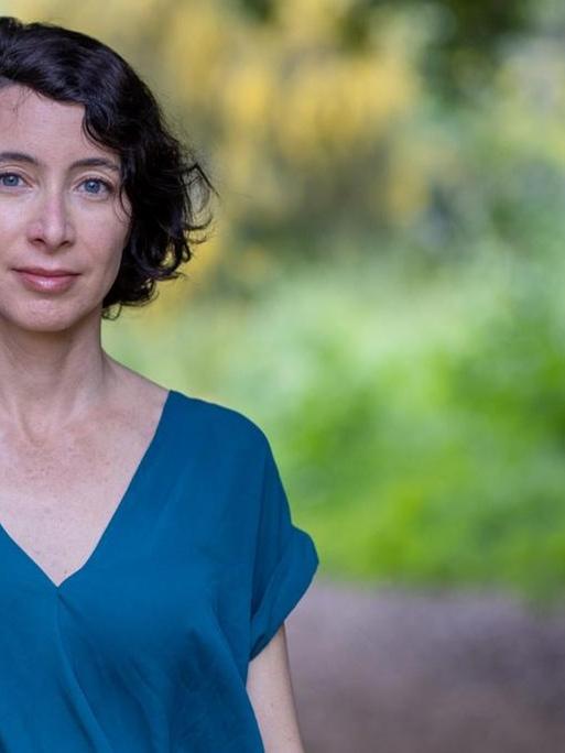 Porträt der Autorin in einem kurzärmeligen, blauen Oberteil unter Bäumen.