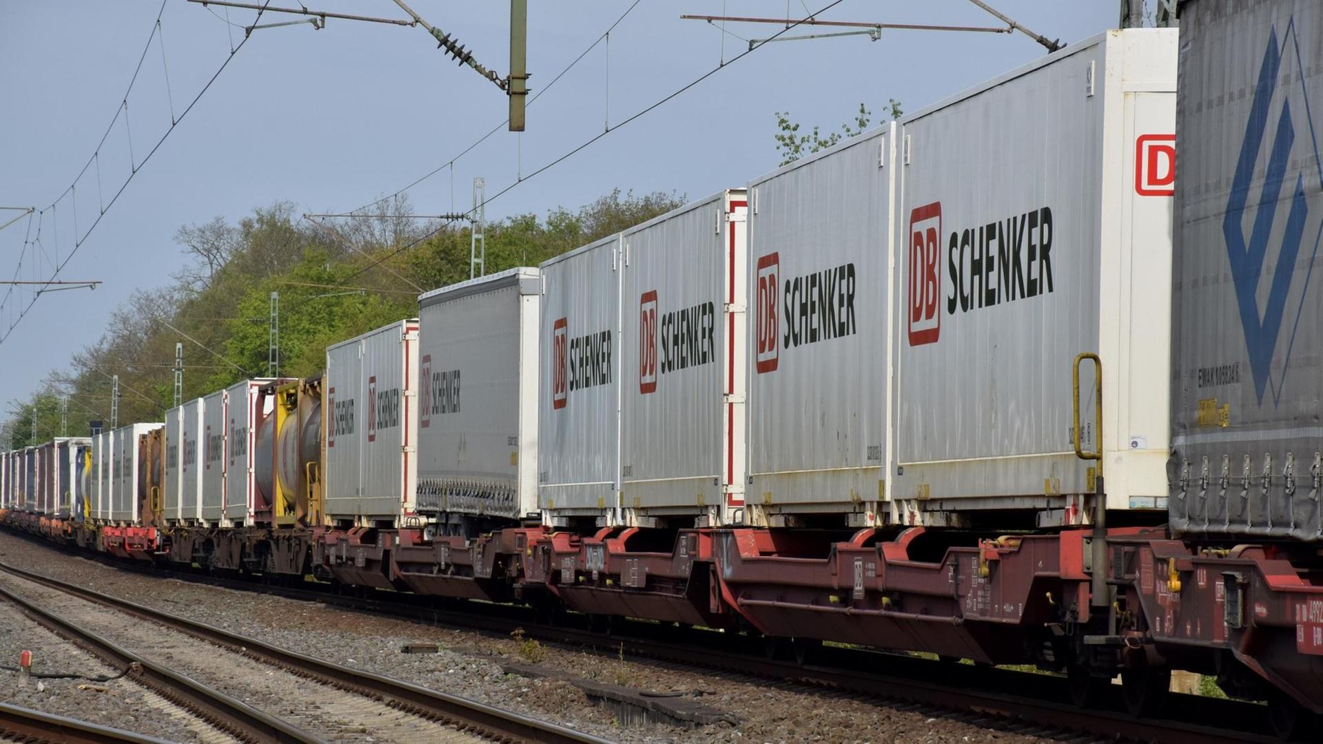 Ein Abschnitt des Zuges ist schräg von der Seite auf einer Schiene fotografiert. Die Container sind weiß und haben die Aufschrift "DB Schenker".