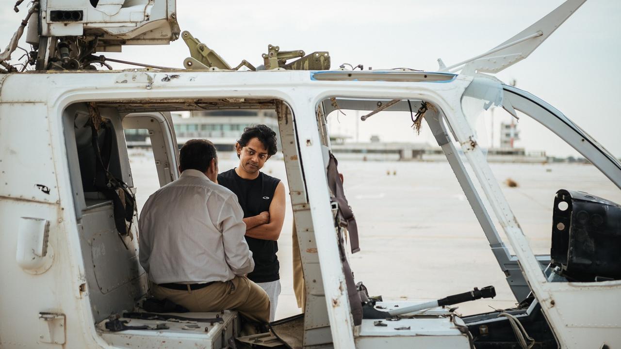 Naeem Mohaiemen steht auf dem verlassenen Flughafen "Athen Ellinikon" vor einem Helikopter, in dem Schauspieler sitzt.