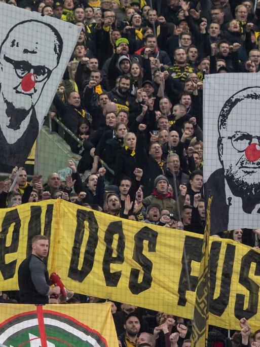 BVB-Fans zeigen Banner mit den Gesichtern von Dietmar Hopp (l-r), Fritz Keller und Karl-Heinz Rummenigge mit roter Clownsnase. Darunter ist ein Banner mit der Aufschrift "Die hässlichen Fratzen des Fußball!".