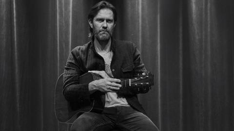 S/w-Bild: Ein Mann mit dunkelblonden längeren Haaren und Bart sitzt auf einem Barhocker, hat eine akustische Gitarre unter den Arm geklemmt. Er blickt direkt in die Kamera.