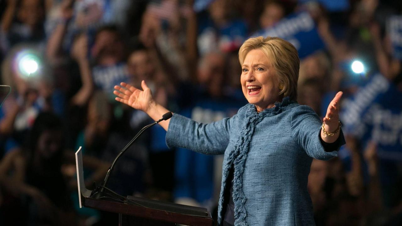 Hillary Clinton vor einem Rednerpult. Sie hat die Arme weit ausgebreitet und strahlt.