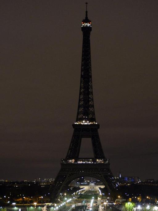 Man sieht den Eiffelturm aus einiger Entfernung in der Nacht. Straßenlaternen leuchten.