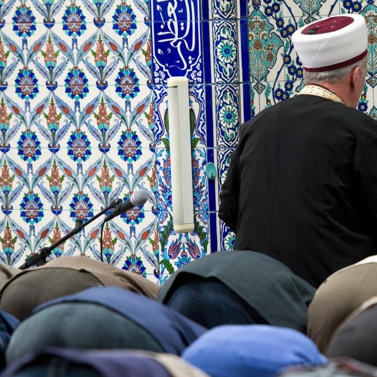 Viele Betende senken den Kopf auf den Boden. Vor ihnen sieht man von hinten den Imam vor einer Wand mit blauen Ornamenten.