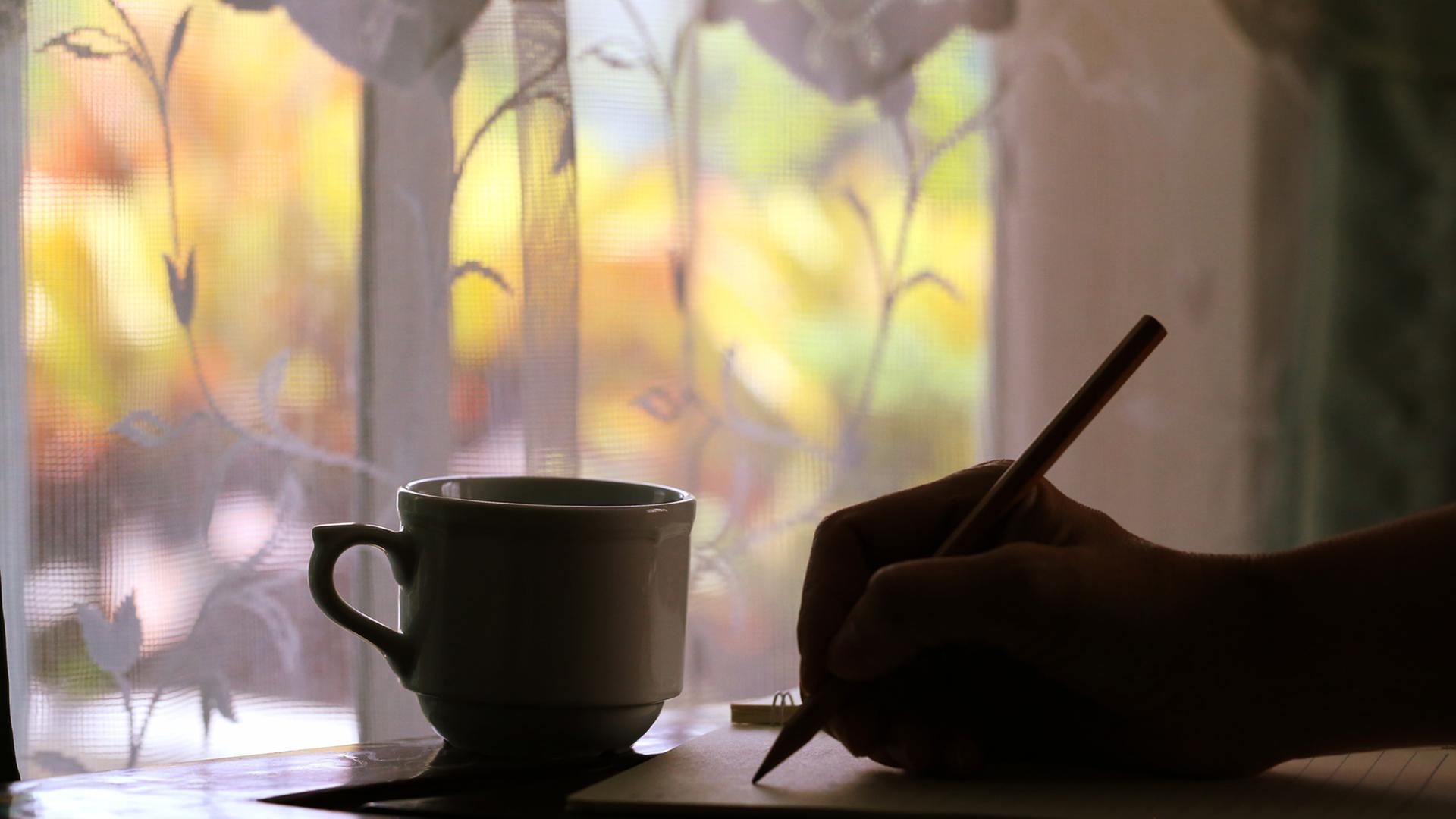 Vor eine, Fenster mit Blick nach draußen steht im Schatten ein Tasse; im Schatten sieht man außerdem eine Hand, die mit einem Stift auf ein Blatt Papier schreibt
