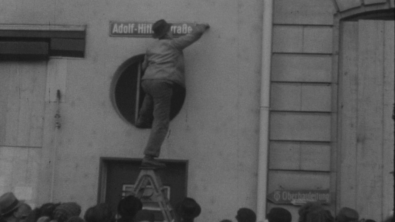 Schwarz-Weiß-Aufnahme eines Mannes, der auf einer Leiter steht und ein Straßenschild mit der Aufschrift "Adolf-Hitler-Straße" abmontiert.