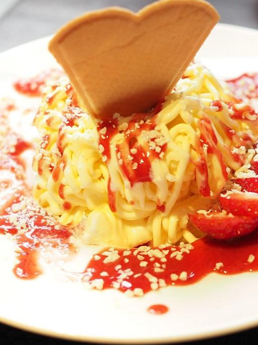Auf einem Teller ist eine Portion Spaghettieis angerichtet. Diese ist mit einer Herzwaffel dekoriert, dazu eine aufgefächerte Erdbeere.