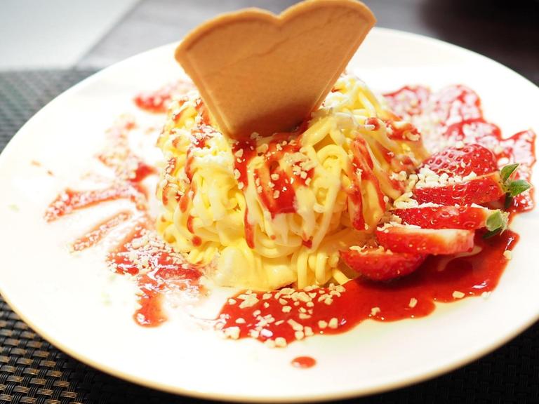 Auf einem Teller ist eine Portion Spaghettieis angerichtet. Diese ist mit einer Herzwaffel dekoriert, dazu eine aufgefächerte Erdbeere.