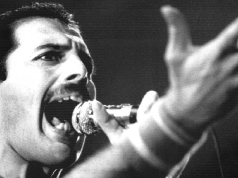 Der Sänger Freddie Mercury sing mit ausgestrecktem Arm in ein Mikrofon