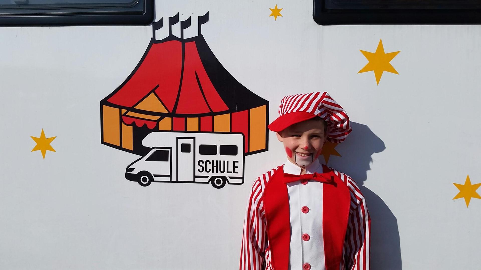 Ein als Clown verkleidetes Kind steht vor einem Campingwagen mit der Aufschrift Schule und Bildern eines Zirkus.