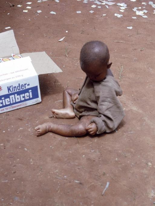 Ein unterernährtes kleines Kind sitzt mit verdecktem Gesicht auf sandigem Boden, daneben eine geöffnete Box, die "Kinder-Grießbrei" der deutschen Marke Aurora beinhaltet.