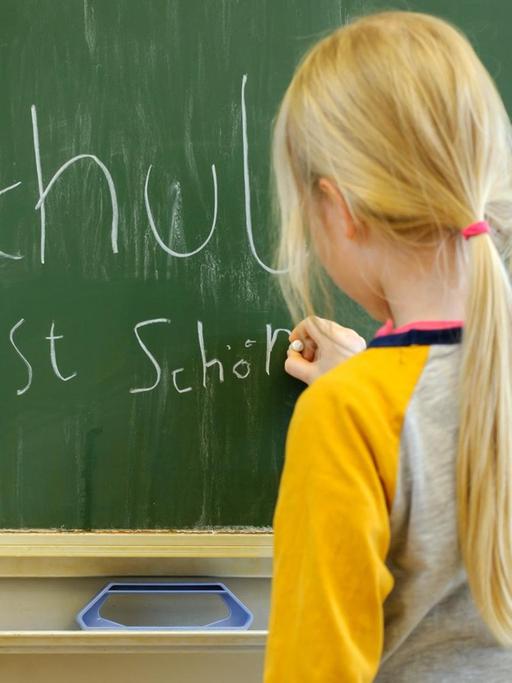 Ein Mädchen schreibt mit Kreide an eine Schultafel den Satz "Schule ist schön".