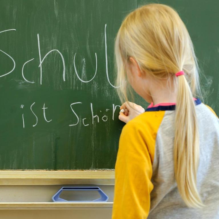 Ein Mädchen schreibt mit Kreide an eine Schultafel den Satz "Schule ist schön".