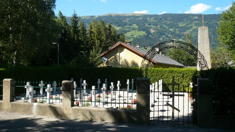 Hinter dem Eingangstor des Friedhofes sind Kreuze zu erkennen. Im Hintergrund bewaldete Berghänge.