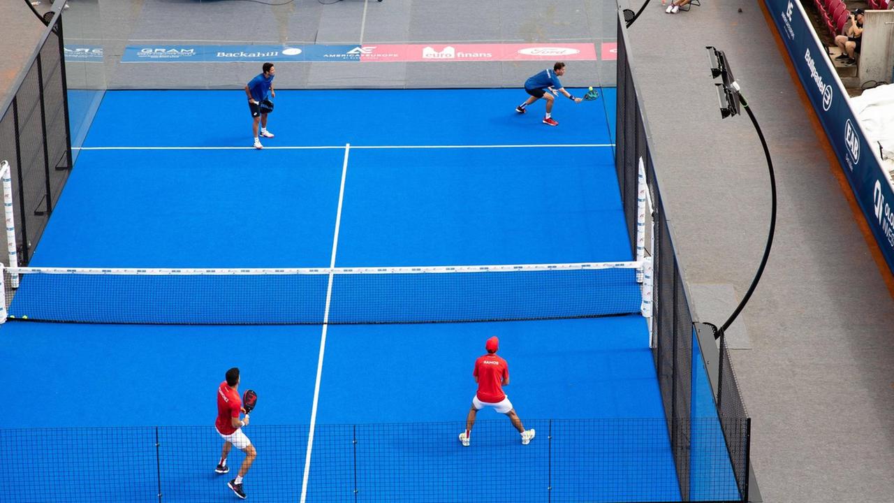 Blick auf einen Padel-Platz mit vier Spielern: Ein Tennis-ähnlicher Platz mit blauem Boden umrahmt von einer transparanten Wand.