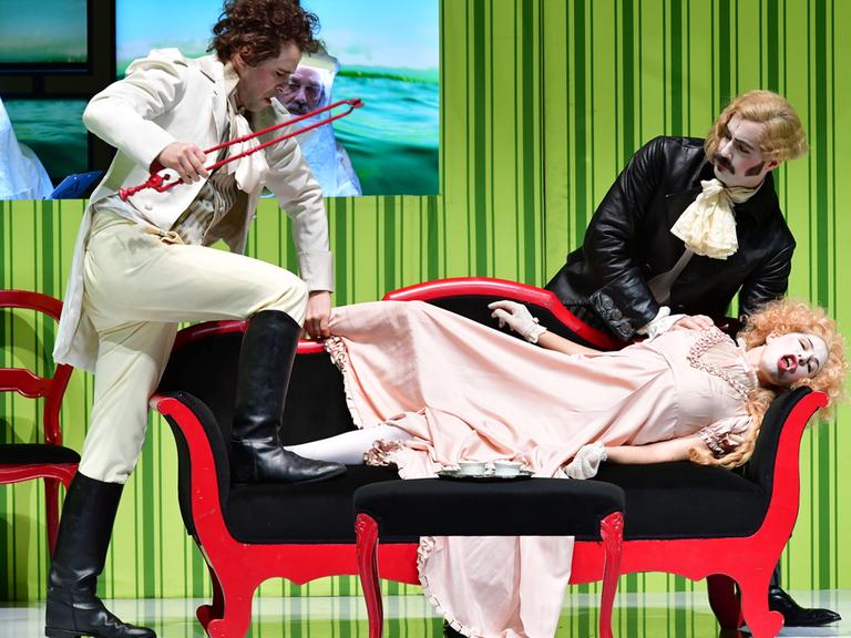 Das Bild zeigt eine Szene aus der Oper "Diodati. Unendlich". Auf einem Sofa liegt eine Frau in Kostüm, zwei Männer stehen neben ihr.