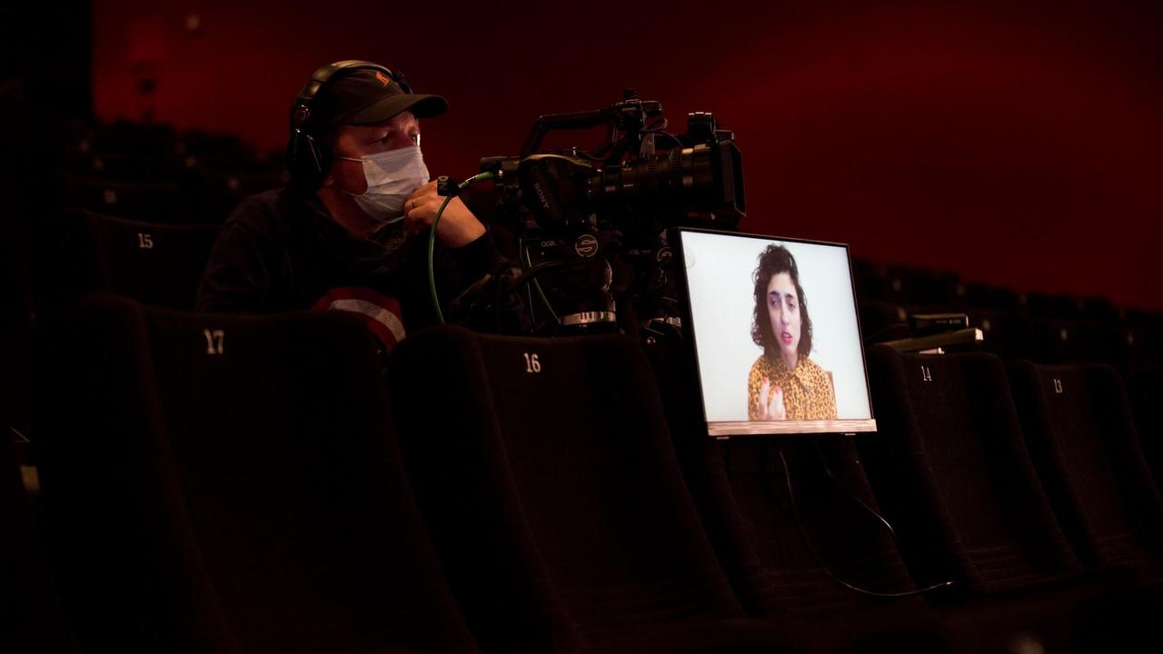 Ein Kameramann mit einer Kamera in einem leeren Kinosaal. Auf einem Sitz steht ein Monitor, auf dem ein Dokumentarfilm gezeigt wird.
