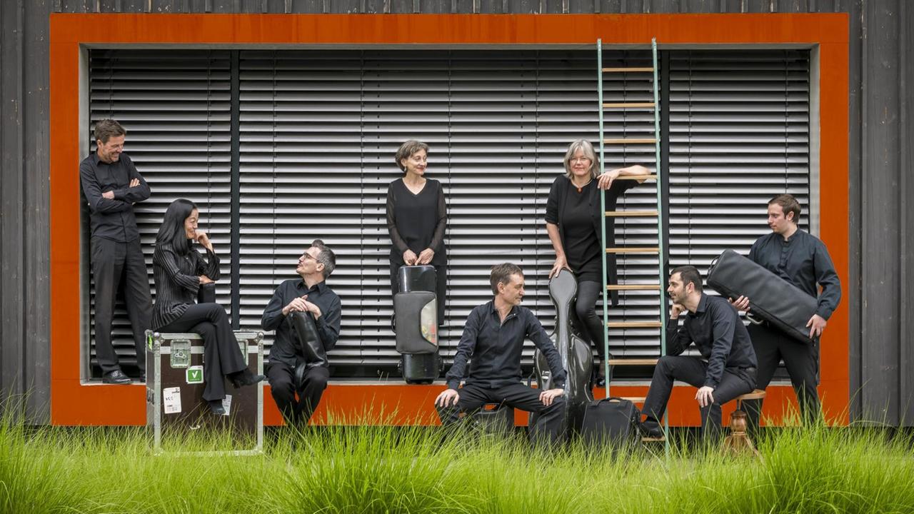 Acht Mitglieder des Ensembles sitzen vor einem Haus mit roten Architekturteilen auf einer Wiese.