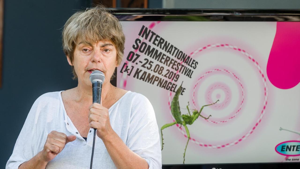 Amelie Deuflhard, Intendantin der Kampnagel-Kulturfabrik ist vor einem Poster des Internationalen Sommerfestivals 2019 mit einem Mikrophon in der Hand zu sehen.