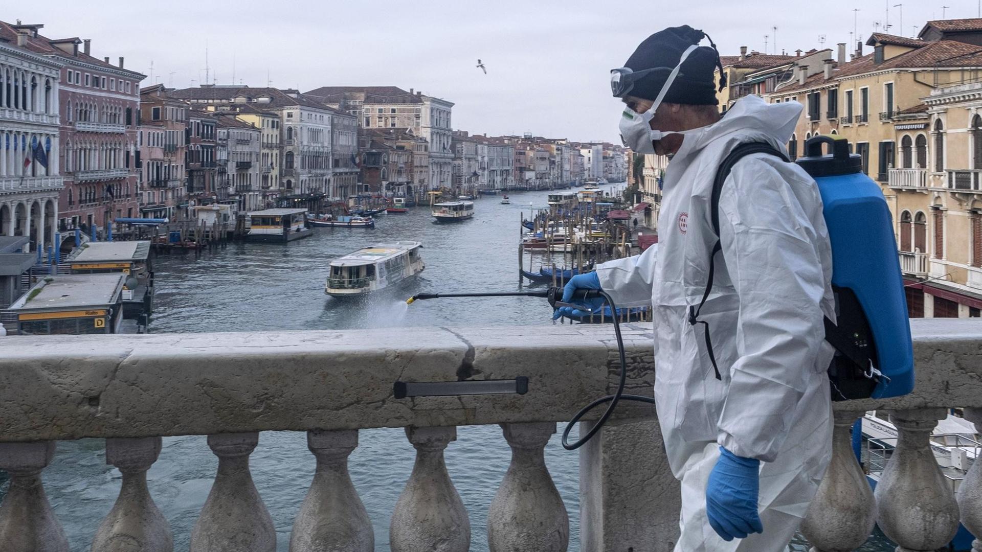 Ein Mann in Schutzanzug desinfiziert den Marmor auf der Rialto-Brücke in Venedig. Im Hintergrund ist der Canal Grande und Häuserfassaden zu sehen.