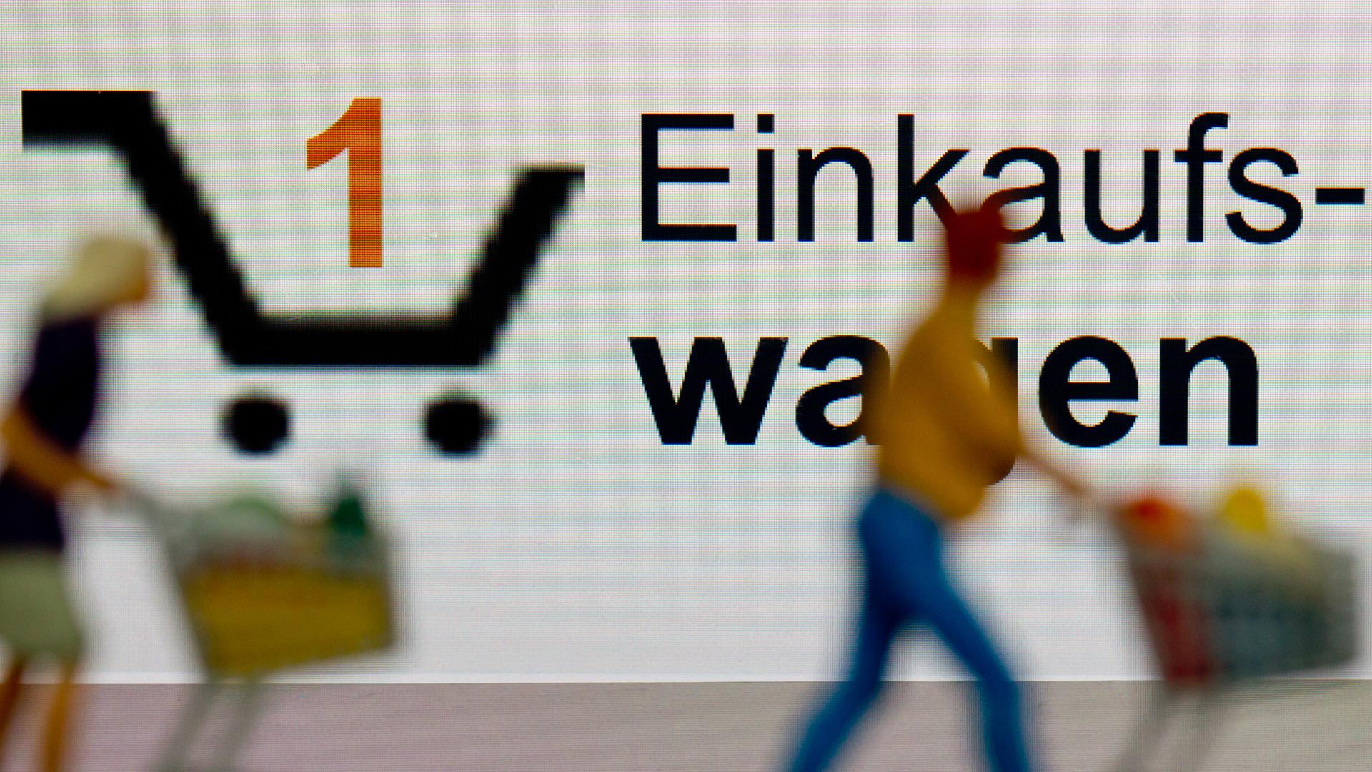 Zwei kleine Plastikfiguren mit Einkaufswagen stehen in Berlin vor einem iPhone auf dem ein virtueller Einkaufswagen dargestellt ist.