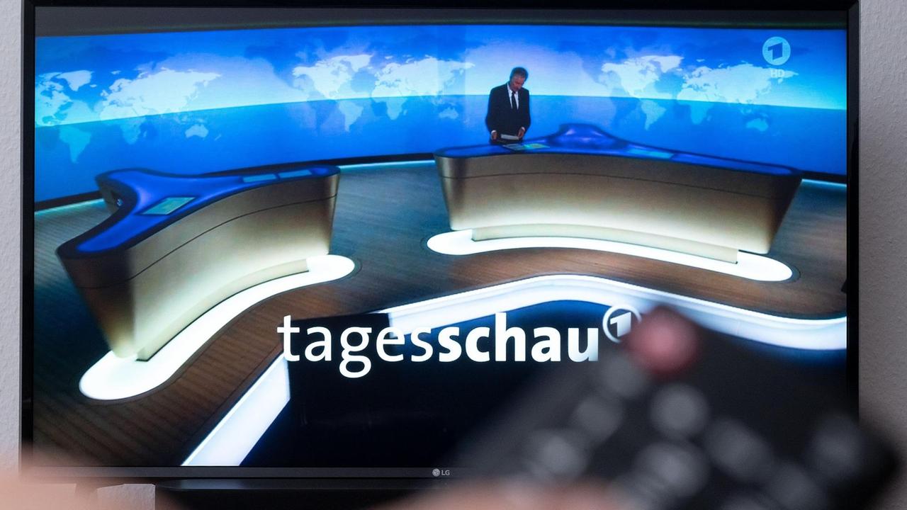 Ein Mann hält eine Fernbedienung vor einen Fernseher, auf dem die Tagesschau läuft.