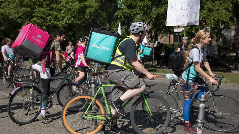 Radfahrer mit pinken und türkisen Liefer-Rucksäcken von Foodora und Deliveroo stehen zusammen an einer Straßenecke.