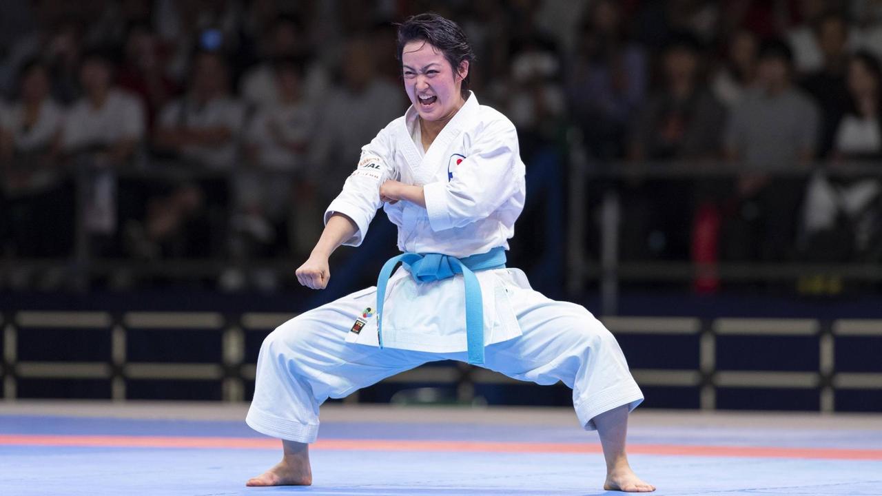 Kiyou Shimizu im Karateanzug mit blauem Gürtel steht breitbeinig in einer Arena, streckt die rechte Faust vor und schreit. Im Hintergrund sind Zuschauer zu sehen.