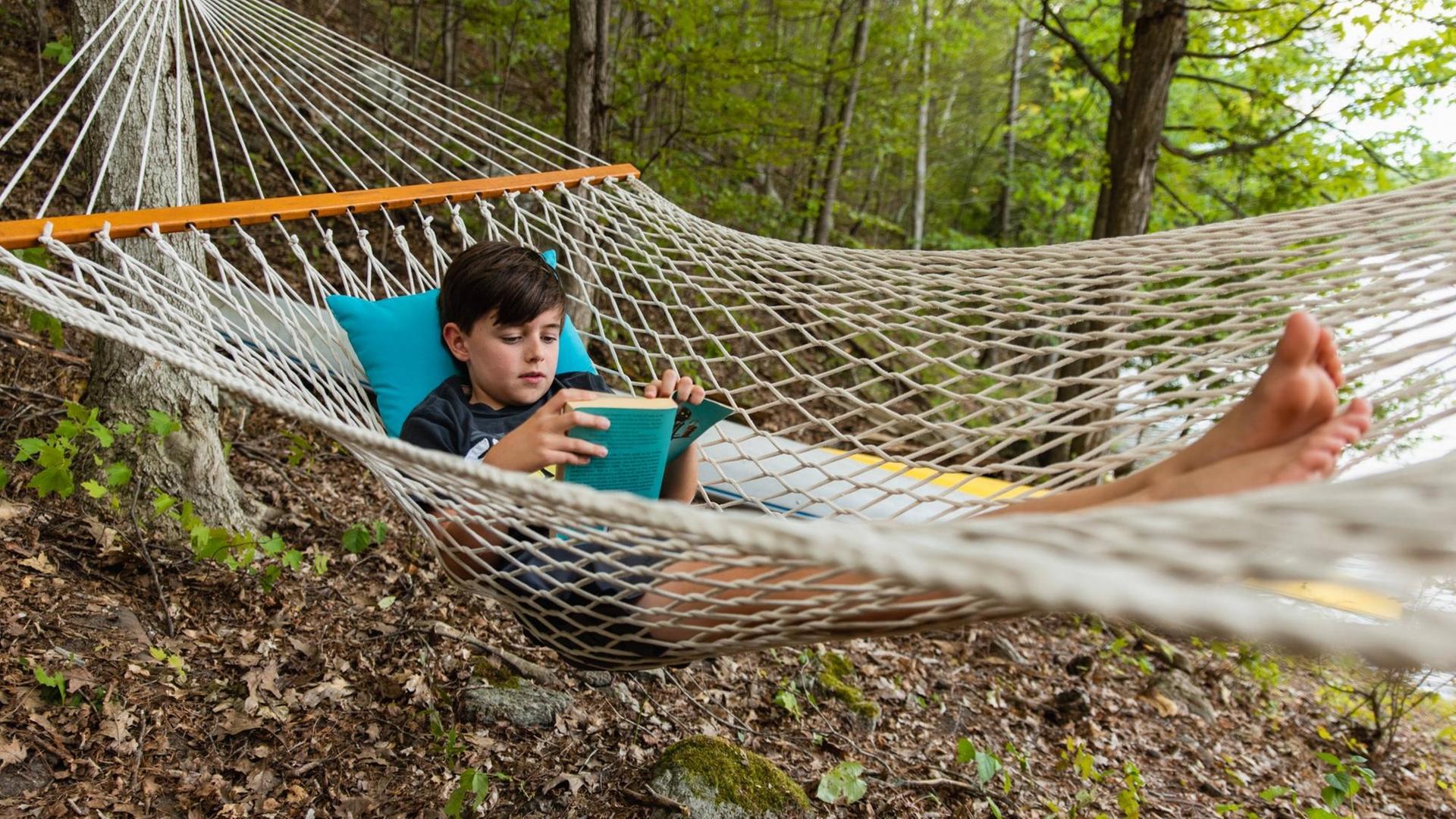 Young boy laying in a woven hammock relaxing and reading a book. Buck Lake, ON, Canada PUBLICATIONxINxGERxSUIxAUTxONLY CR_DOHO200904J-485083-01 ,model released, Symbolfoto