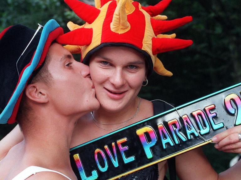 Mit einem Schild "Love Parade 97" in der Hand demonstriert ein Pärchen, mit Hüten bunt geschmückt, den Sinn des Festes - und gibt sich einen Kuss. Fast eine Million Techno-Fans haben am 12.07.1997 bei der Love Parade in Berlin die weltweit größte Party des Jahres gefeiert.