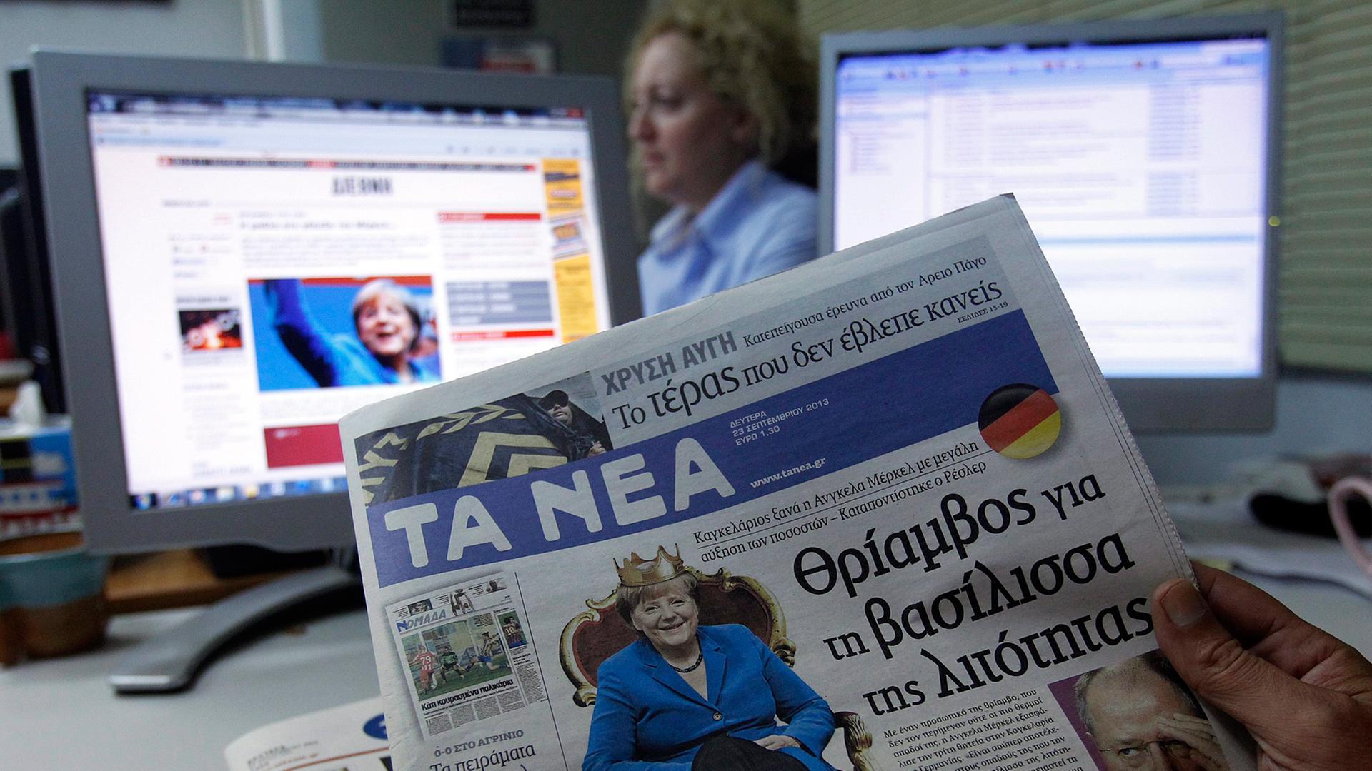 Ein griechischer Journalist hält eine Tageszeitung in der Hand, auf der Bundeskanzlerin Merkel auf einem Thron sitzt.