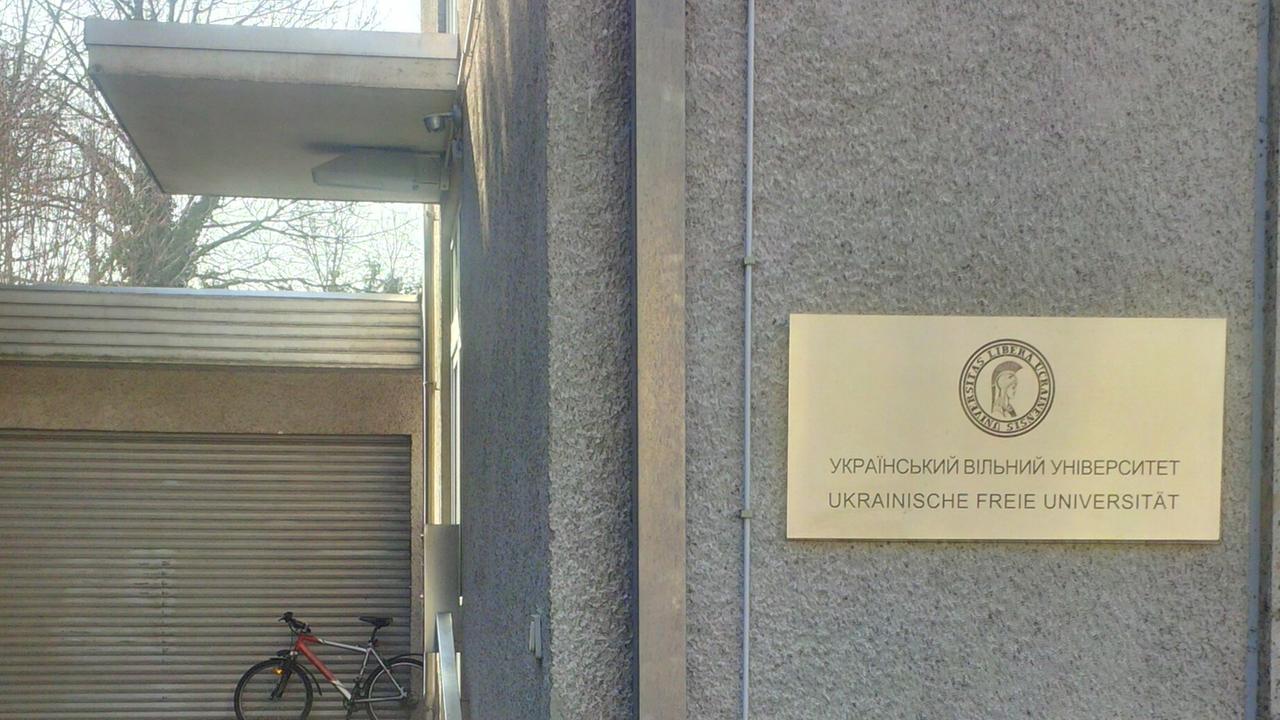 Goldenes Schild mit Aufschrift "Ukrainische Freie Universität München", auch in Kyrillisch an einer grauen Hauswand, im Hintergrund steht ein Fahrrad an einer Wand.
